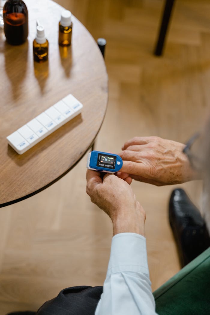 An elderly individual checks blood oxygen using a pulse oximeter beside a table with medicine bottles.